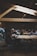 A dimly lit coffee shop features wooden beams on the ceiling and large communal tables. People are sitting on stools, engaging in conversations, working on laptops, and sipping drinks. The atmosphere suggests a casual, modern setting with a focus on both work and social interaction.