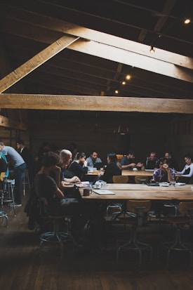 A dimly lit coffee shop features wooden beams on the ceiling and large communal tables. People are sitting on stools, engaging in conversations, working on laptops, and sipping drinks. The atmosphere suggests a casual, modern setting with a focus on both work and social interaction.