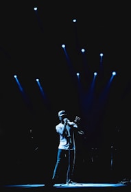 A performer stands on stage under multiple blue spotlights, holding a microphone and wearing a cap and casual clothes. The backdrop is dark, highlighting the illuminated area around the performer.