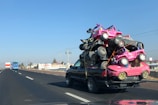 A pickup truck is traveling on a highway with a large load of colorful toy cars stacked on its bed. The cars are primarily pink and tied down with orange straps. In the background, the road stretches ahead with a few other vehicles visible, including a blue bus. The sky is clear and blue, and there are electric poles lined along the side of the road.
