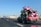 A pickup truck is traveling on a highway with a large load of colorful toy cars stacked on its bed. The cars are primarily pink and tied down with orange straps. In the background, the road stretches ahead with a few other vehicles visible, including a blue bus. The sky is clear and blue, and there are electric poles lined along the side of the road.
