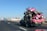 A pickup truck is traveling on a highway with a large load of colorful toy cars stacked on its bed. The cars are primarily pink and tied down with orange straps. In the background, the road stretches ahead with a few other vehicles visible, including a blue bus. The sky is clear and blue, and there are electric poles lined along the side of the road.