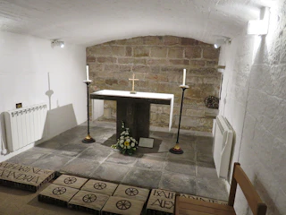 A small, intimate stone chapel with a simple altar featuring two white candles and a small cross. The space is enclosed with white-washed brick walls and a brick archway ceiling, furnished with minimalistic décor including a bouquet of flowers. The floor has stone tiles and inscribed plaques.