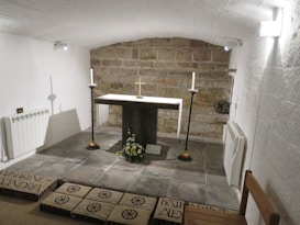 A small, intimate stone chapel with a simple altar featuring two white candles and a small cross. The space is enclosed with white-washed brick walls and a brick archway ceiling, furnished with minimalistic d&eacute;cor including a bouquet of flowers. The floor has stone tiles and inscribed plaques.