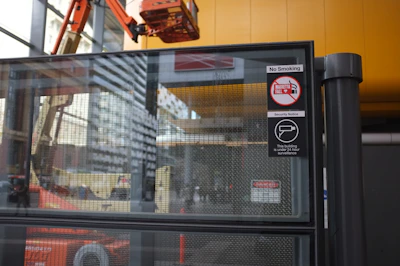 Glass partition with mesh and signs indicating no smoking and 24-hour surveillance. A construction lift is visible in the background inside a modern building with yellow and grey elements.