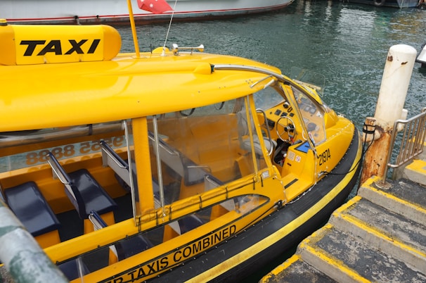 A bright yellow water taxi is docked beside a pier with steps leading down to the water. The taxi has clear windows and seating inside, with a steering wheel visible in the cabin area. Part of a water channel is in the background, along with another boat.