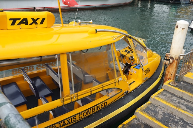 A bright yellow water taxi is docked beside a pier with steps leading down to the water. The taxi has clear windows and seating inside, with a steering wheel visible in the cabin area. Part of a water channel is in the background, along with another boat.