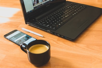 A black laptop sits open on a wooden surface, next to a smartphone displaying a website. A white pen is placed beside the phone, and a black mug filled with coffee is in the foreground.