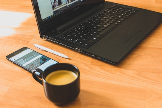 A black laptop sits open on a wooden surface, next to a smartphone displaying a website. A white pen is placed beside the phone, and a black mug filled with coffee is in the foreground.