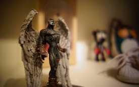 A detailed figurine of a powerful, muscular character with large, intricate wings, wearing dark armor with metallic accents, stands prominently in the foreground. The background is softly blurred, featuring dimly visible objects including another figurine and a plush toy.