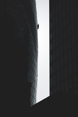 A person is suspended on ropes, cleaning or performing maintenance work on the exterior of a tall, modern building with a distinctive curved and glass-paneled architecture. The structure's surface contrasts with the silhouette of adjacent buildings, emphasizing the height and scale.