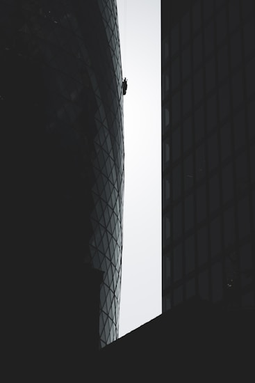 A person is suspended on ropes, cleaning or performing maintenance work on the exterior of a tall, modern building with a distinctive curved and glass-paneled architecture. The structure's surface contrasts with the silhouette of adjacent buildings, emphasizing the height and scale.
