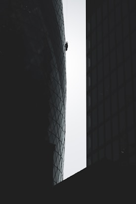 A person is suspended on ropes, cleaning or performing maintenance work on the exterior of a tall, modern building with a distinctive curved and glass-paneled architecture. The structure's surface contrasts with the silhouette of adjacent buildings, emphasizing the height and scale.