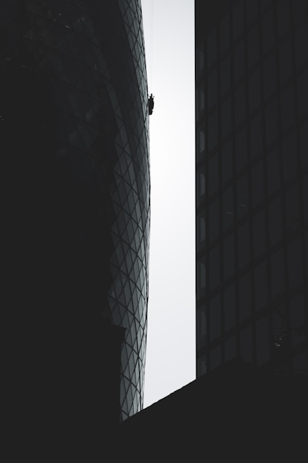 A person is suspended on ropes, cleaning or performing maintenance work on the exterior of a tall, modern building with a distinctive curved and glass-paneled architecture. The structure's surface contrasts with the silhouette of adjacent buildings, emphasizing the height and scale.
