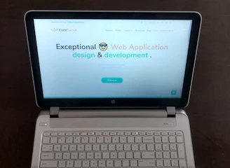 A laptop computer with a website displayed on the screen featuring the text 'Exceptional Web Application design & development'. The laptop is on a dark surface, and the website has a clean and modern design with a light background and colorful text elements.