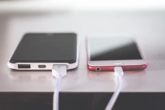 Two smartphones are placed side by side on a flat surface, each connected to a charging cable. The phone on the left is encased in a black and white case, while the one on the right has a pink hue. Both phones appear to be charging simultaneously, with the cables extending out of sight.