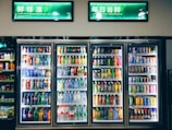 Refrigerated display cases contain a wide assortment of colorful beverages, including bottles and cans of different sizes and shapes. Shelves are neatly organized, featuring various drinks like water, soda, juice, and energy drinks. Bright lighting highlights the vivid packaging and labels.