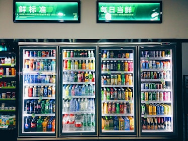 Refrigerated display cases contain a wide assortment of colorful beverages, including bottles and cans of different sizes and shapes. Shelves are neatly organized, featuring various drinks like water, soda, juice, and energy drinks. Bright lighting highlights the vivid packaging and labels.