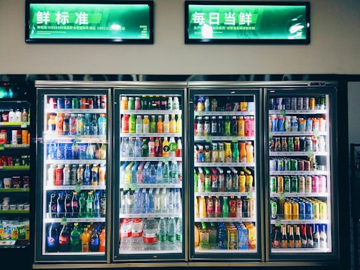Refrigerated display cases contain a wide assortment of colorful beverages, including bottles and cans of different sizes and shapes. Shelves are neatly organized, featuring various drinks like water, soda, juice, and energy drinks. Bright lighting highlights the vivid packaging and labels.