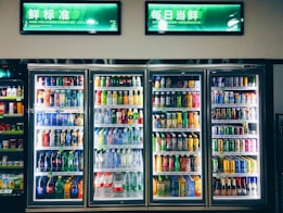 Refrigerated display cases contain a wide assortment of colorful beverages, including bottles and cans of different sizes and shapes. Shelves are neatly organized, featuring various drinks like water, soda, juice, and energy drinks. Bright lighting highlights the vivid packaging and labels.