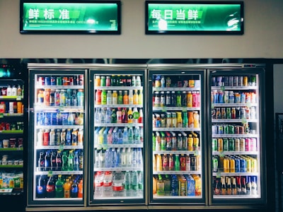 Refrigerated display cases contain a wide assortment of colorful beverages, including bottles and cans of different sizes and shapes. Shelves are neatly organized, featuring various drinks like water, soda, juice, and energy drinks. Bright lighting highlights the vivid packaging and labels.