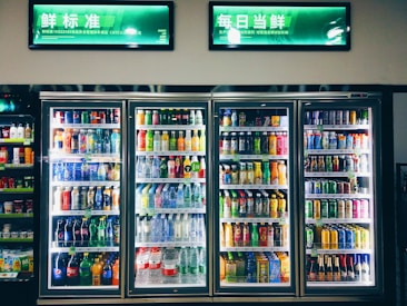 Refrigerated display cases contain a wide assortment of colorful beverages, including bottles and cans of different sizes and shapes. Shelves are neatly organized, featuring various drinks like water, soda, juice, and energy drinks. Bright lighting highlights the vivid packaging and labels.
