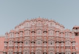 An ornate red and pink sandstone palace facade featuring a series of intricately designed windows with white latticework and arches. The architecture is symmetrical and grand, exhibiting traditional elements of Indian design against a clear blue sky.
