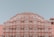 An ornate red and pink sandstone palace facade featuring a series of intricately designed windows with white latticework and arches. The architecture is symmetrical and grand, exhibiting traditional elements of Indian design against a clear blue sky.
