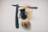 A straight razor, shaving brush, and a shaving cream bowl are arranged closely together on a light background. The straight razor has a black handle and a shiny metal blade. The shaving brush features a black handle and light bristles, resting against the side of the cream-colored bowl.