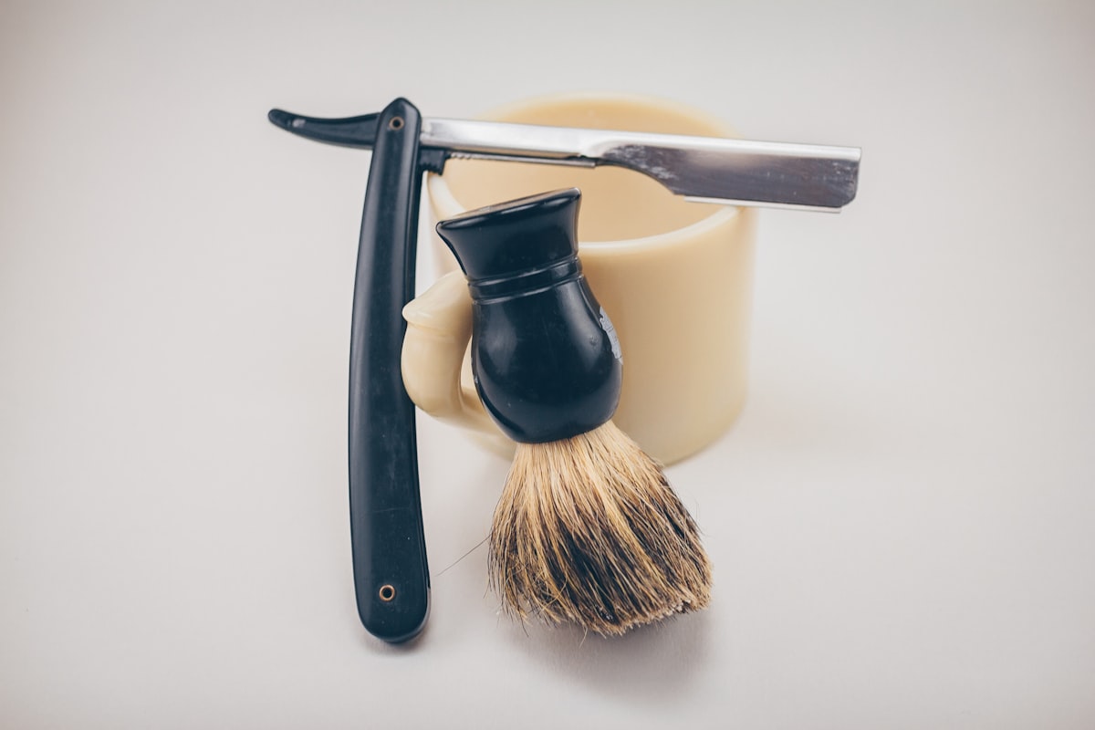 A straight razor resting beside a ceramic mug and a shaving brush