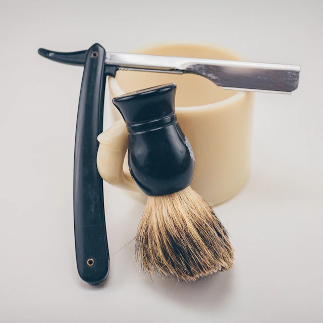 A black straight razor beside a beige ceramic mug and a shaving brush