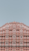 An intricate pink sandstone building featuring numerous ornate windows and arched designs. The architecture is symmetrical, showcasing detailed carvings and a historic style against a clear blue sky.