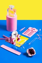 A flat lay composition featuring an assortment of beauty and fashion items arranged on a bright blue surface. A pink pedestal holds a glass perfume bottle, while a mirror reflects a pair of pink and yellow eyeball stress balls. Nearby, a pink sheet with false eyelashes, a pair of tweezers, scissors, and a hair clip are placed. A pair of clear glasses, a pink comb, and a geode rock complete the eclectic assembly. The vivid yellow and blue background adds to the vibrant and quirky feel.