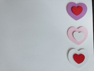 Three pairs of layered hearts are arranged vertically on the right, each consisting of an outer and inner heart. The colors of the hearts from top to bottom are purple with red, pink with white, and white with red. The background is a plain light gray or white surface.