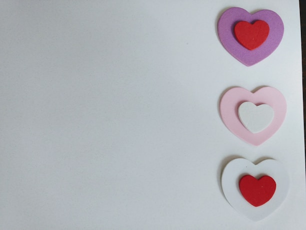Three pairs of layered hearts are arranged vertically on the right, each consisting of an outer and inner heart. The colors of the hearts from top to bottom are purple with red, pink with white, and white with red. The background is a plain light gray or white surface.