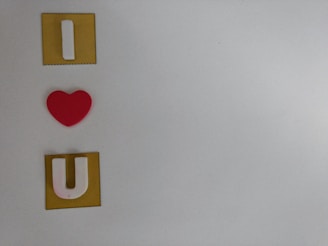 The image features three elements vertically aligned against a gray background. At the top is a white letter 'I' on a yellow square, followed by a red heart shape positioned centrally, and at the bottom is a white letter 'U' on another yellow square.