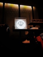 A glowing neon smiley face is centrally placed on a shelf, creating a focal point against a textured wall with dim industrial lighting. Nearby are neatly folded clothes, a white sneaker, and various apparel items, suggesting a retail environment.