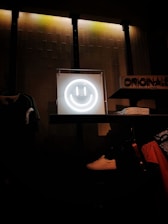 A glowing neon smiley face is centrally placed on a shelf, creating a focal point against a textured wall with dim industrial lighting. Nearby are neatly folded clothes, a white sneaker, and various apparel items, suggesting a retail environment.