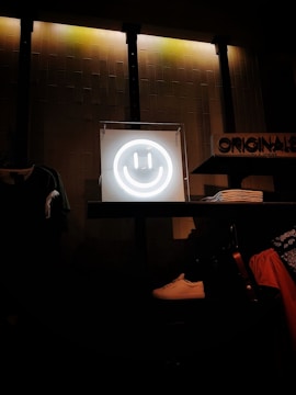 A glowing neon smiley face is centrally placed on a shelf, creating a focal point against a textured wall with dim industrial lighting. Nearby are neatly folded clothes, a white sneaker, and various apparel items, suggesting a retail environment.
