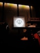 A glowing neon smiley face is centrally placed on a shelf, creating a focal point against a textured wall with dim industrial lighting. Nearby are neatly folded clothes, a white sneaker, and various apparel items, suggesting a retail environment.