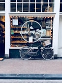 A storefront of a bakery named 'Bake my Day' is visible with a vintage bicycle parked in front. The window display is filled with baked goods and illuminated by warmly lit string lights inside. Various signs and menu boards can be seen through the window, advertising coffee and sandwiches.