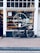 A storefront of a bakery named 'Bake my Day' is visible with a vintage bicycle parked in front. The window display is filled with baked goods and illuminated by warmly lit string lights inside. Various signs and menu boards can be seen through the window, advertising coffee and sandwiches.