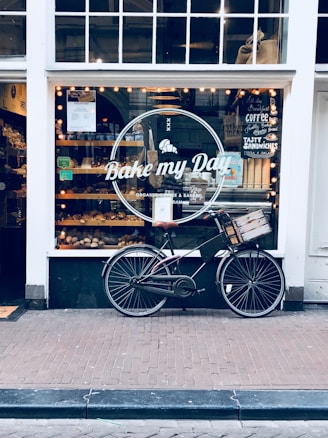 A storefront of a bakery named 'Bake my Day' is visible with a vintage bicycle parked in front. The window display is filled with baked goods and illuminated by warmly lit string lights inside. Various signs and menu boards can be seen through the window, advertising coffee and sandwiches.