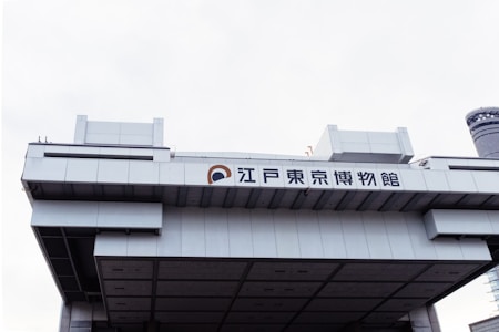 A large, modern, architectural building with Japanese characters displayed prominently on the facade. The structure features a geometric design with clean lines and a metallic texture. The sky is overcast, enhancing the industrial appearance of the building.