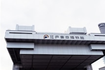 A large, modern, architectural building with Japanese characters displayed prominently on the facade. The structure features a geometric design with clean lines and a metallic texture. The sky is overcast, enhancing the industrial appearance of the building.