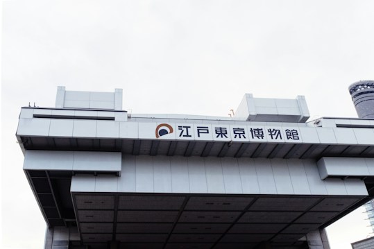 A large, modern, architectural building with Japanese characters displayed prominently on the facade. The structure features a geometric design with clean lines and a metallic texture. The sky is overcast, enhancing the industrial appearance of the building.