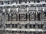 Intricate stone carvings depicting various figures and scenes, likely from mythology, adorn the wall of an ancient structure. The carvings are highly detailed with numerous human and possibly divine figures arranged in tiers.