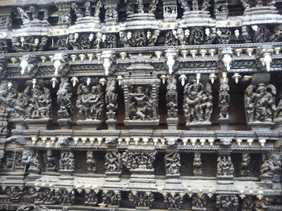 Intricate stone carvings depicting various figures and scenes, likely from mythology, adorn the wall of an ancient structure. The carvings are highly detailed with numerous human and possibly divine figures arranged in tiers.