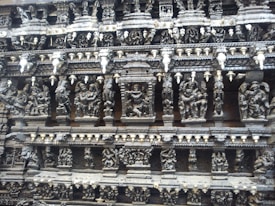 Intricate stone carvings depicting various figures and scenes, likely from mythology, adorn the wall of an ancient structure. The carvings are highly detailed with numerous human and possibly divine figures arranged in tiers.