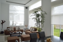 A contemporary living room with large windows allowing natural light. There's a brown modular sofa with patterned cushions, a black coffee table with decorative objects, and a tall indoor plant. A modern sculpture stands near a window. The adjacent dining area has a dark table with candles as the centerpiece.